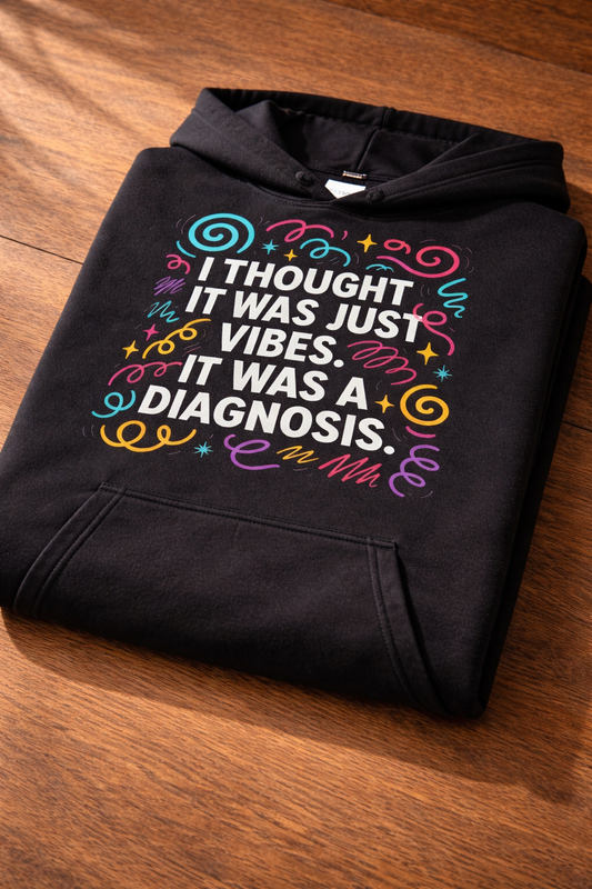 Folded black hoodie on a wooden surface, showing a close-up of the colourful “I thought it was just vibes. It was a diagnosis” design and print texture.