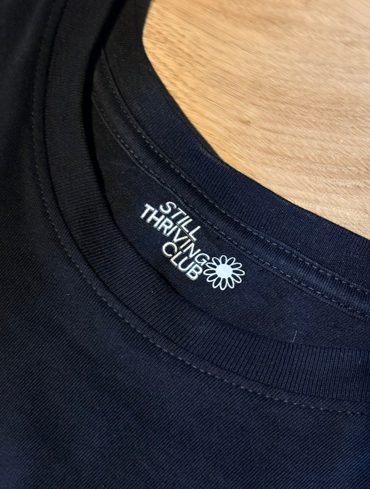 Close-up of a black t-shirt neckline showing no tags just a printed 'Still Thriving Club' logo on a wooden surface