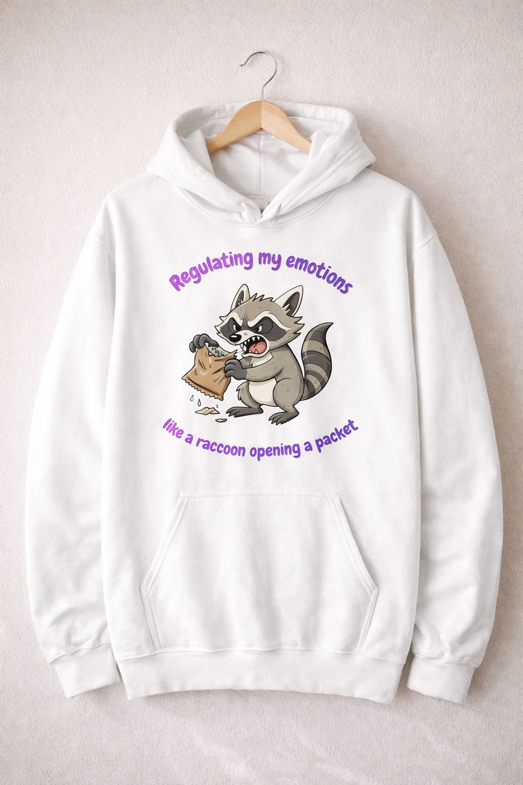 White hoodie on a wooden hanger featuring a cartoon raccoon and the text “Regulating my emotions like a raccoon opening a packet”