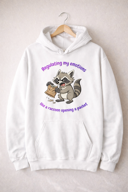 White hoodie on a wooden hanger featuring a cartoon raccoon and the text “Regulating my emotions like a raccoon opening a packet”