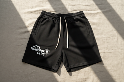 Black unisex cotton shorts with drawstring waist and Still Thriving Club logo on the left leg, laid flat on neutral fabric background