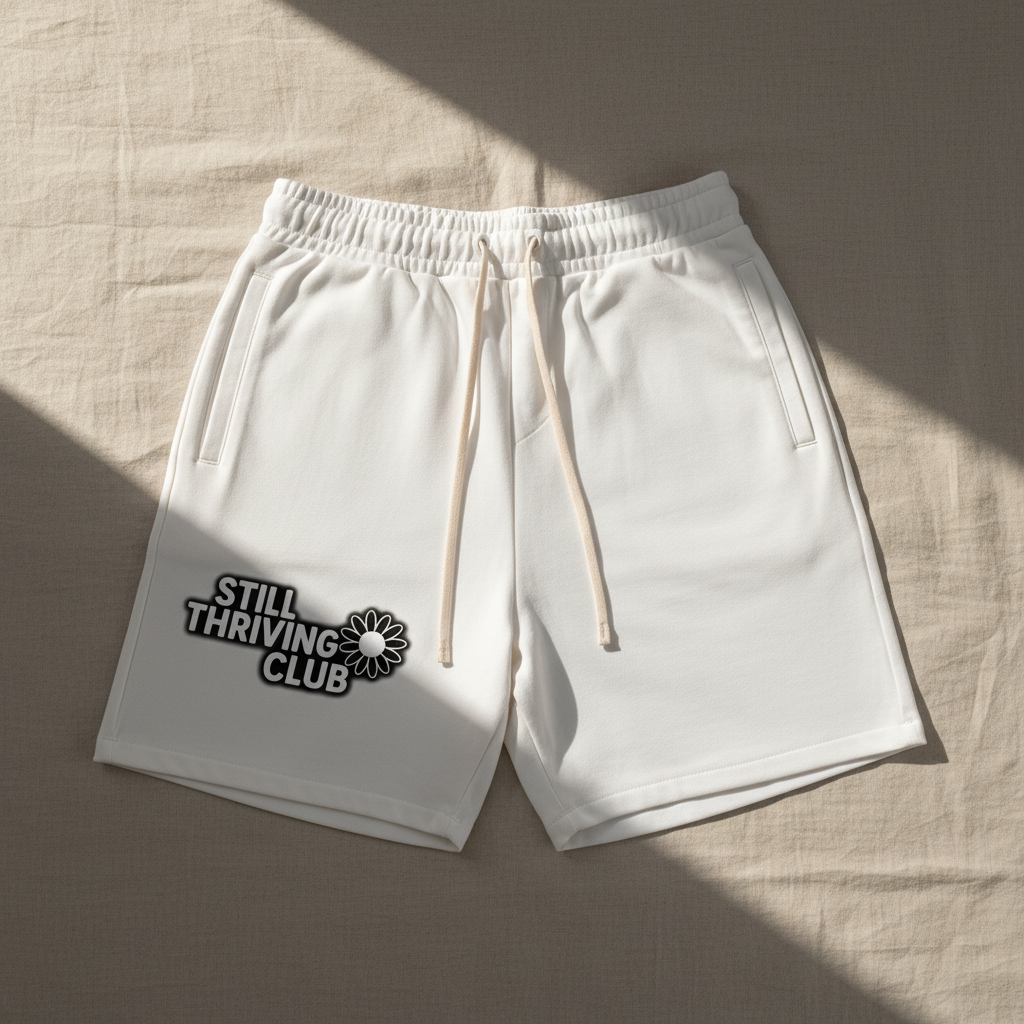 White unisex cotton shorts with adjustable drawstring and Still Thriving Club logo on the left leg, laid flat in soft natural lighting