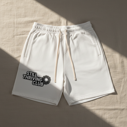 White unisex cotton shorts with adjustable drawstring and Still Thriving Club logo on the left leg, laid flat in soft natural lighting
