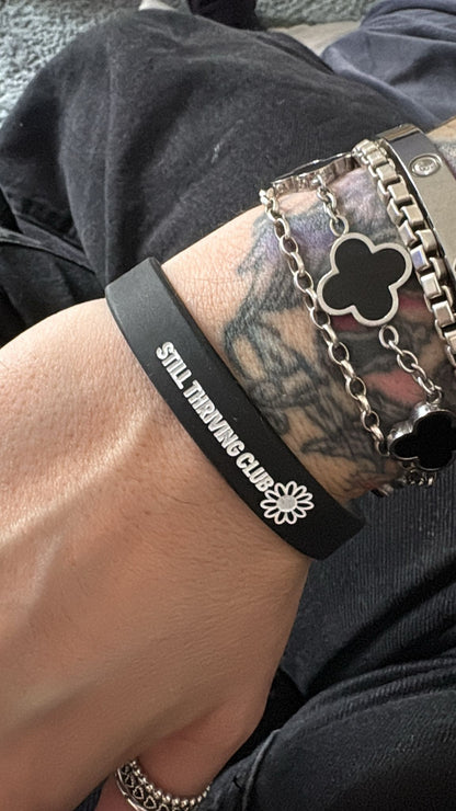 Black Still Thriving Club wristband worn on a wrist alongside silver bracelets, showing the logo and flower detail up close.