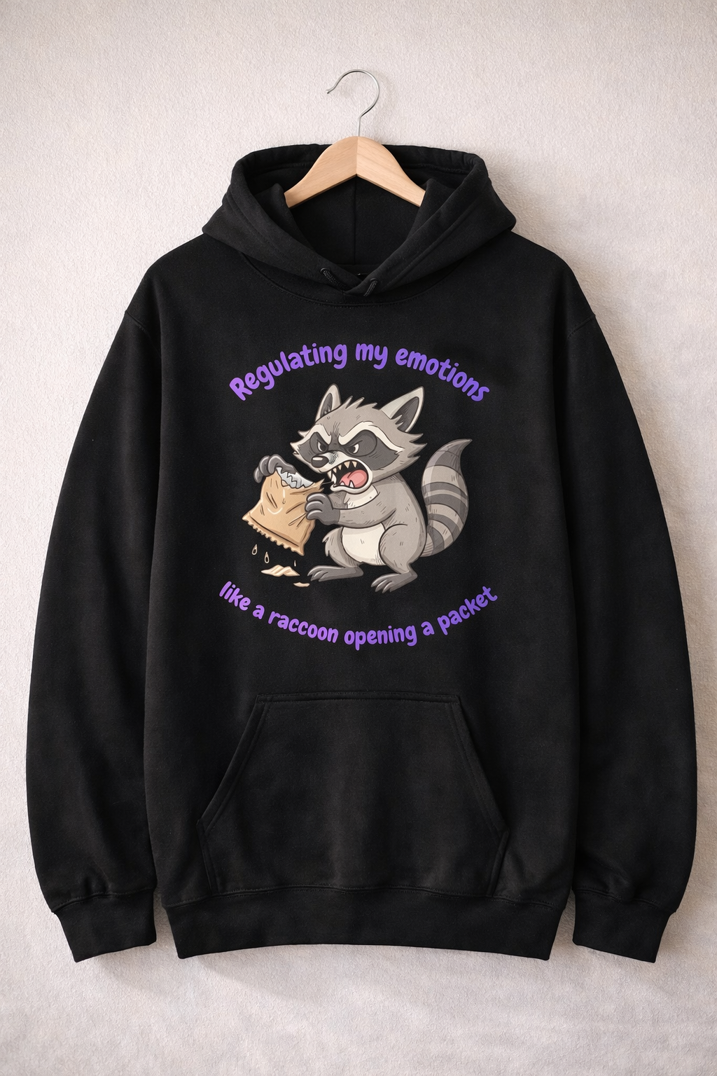 Black hoodie on a wooden hanger with a raccoon illustration and the phrase “Regulating my emotions like a raccoon opening a packet”