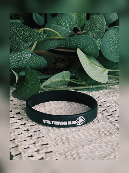 Black Still Thriving Club silicone wristband resting flat on a textured surface with leafy plants in the background.