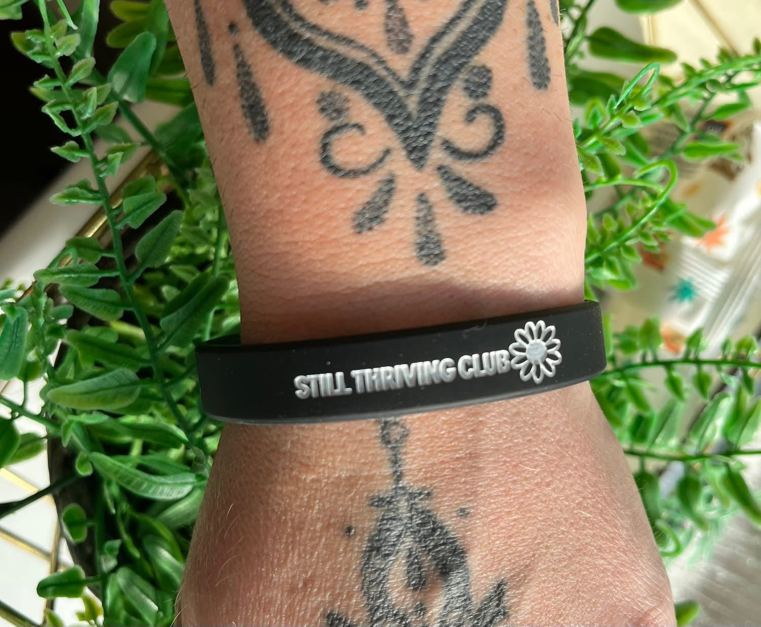 Black silicone Still Thriving Club wristband worn on a tattooed wrist, photographed against green leaves in natural light.