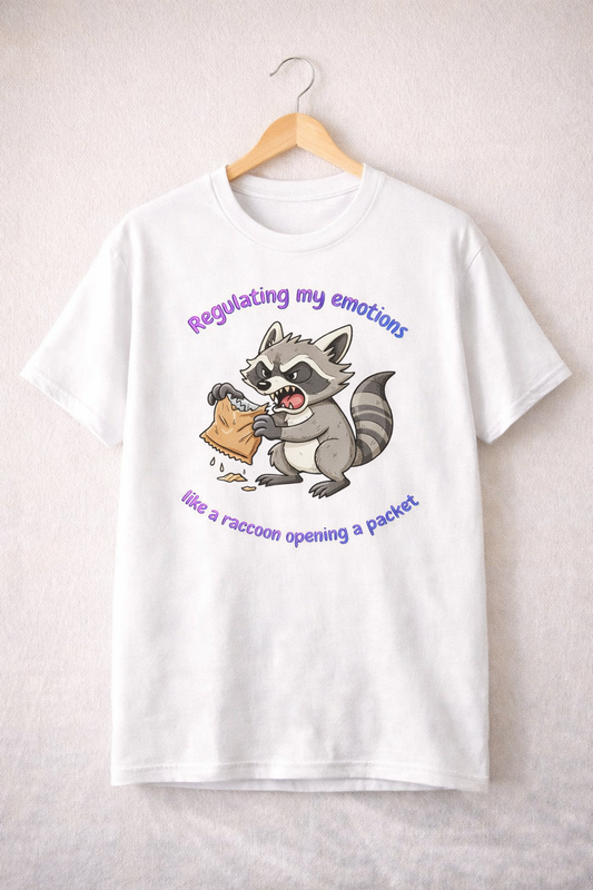 White unisex T-shirt hanging on a wooden hanger with a raccoon illustration and the text “Regulating my emotions like a raccoon opening a packet”