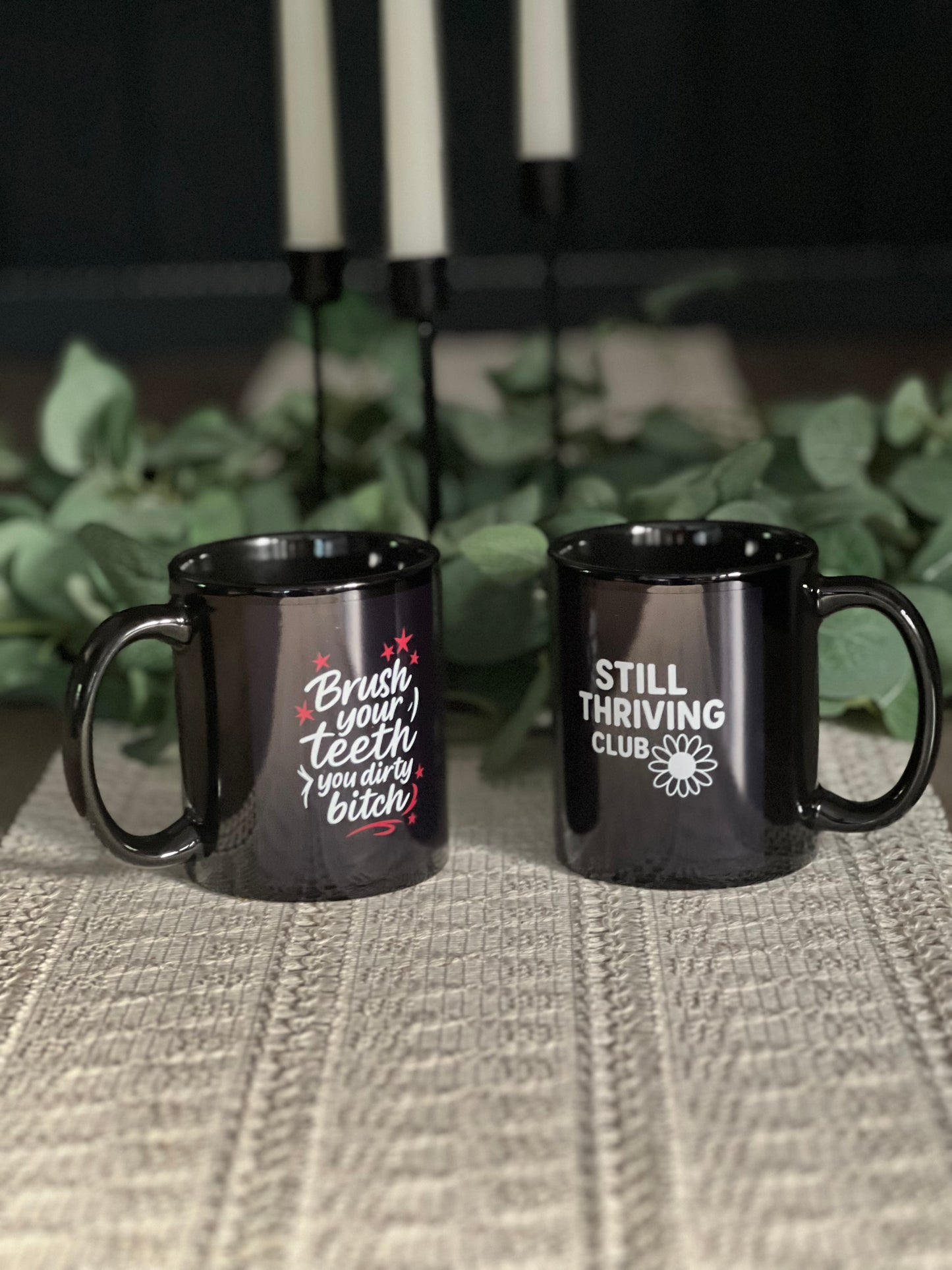 Two black ceramic mugs with Still Thriving Club designs placed on a textured surface, with green plants softly blurred in the background.