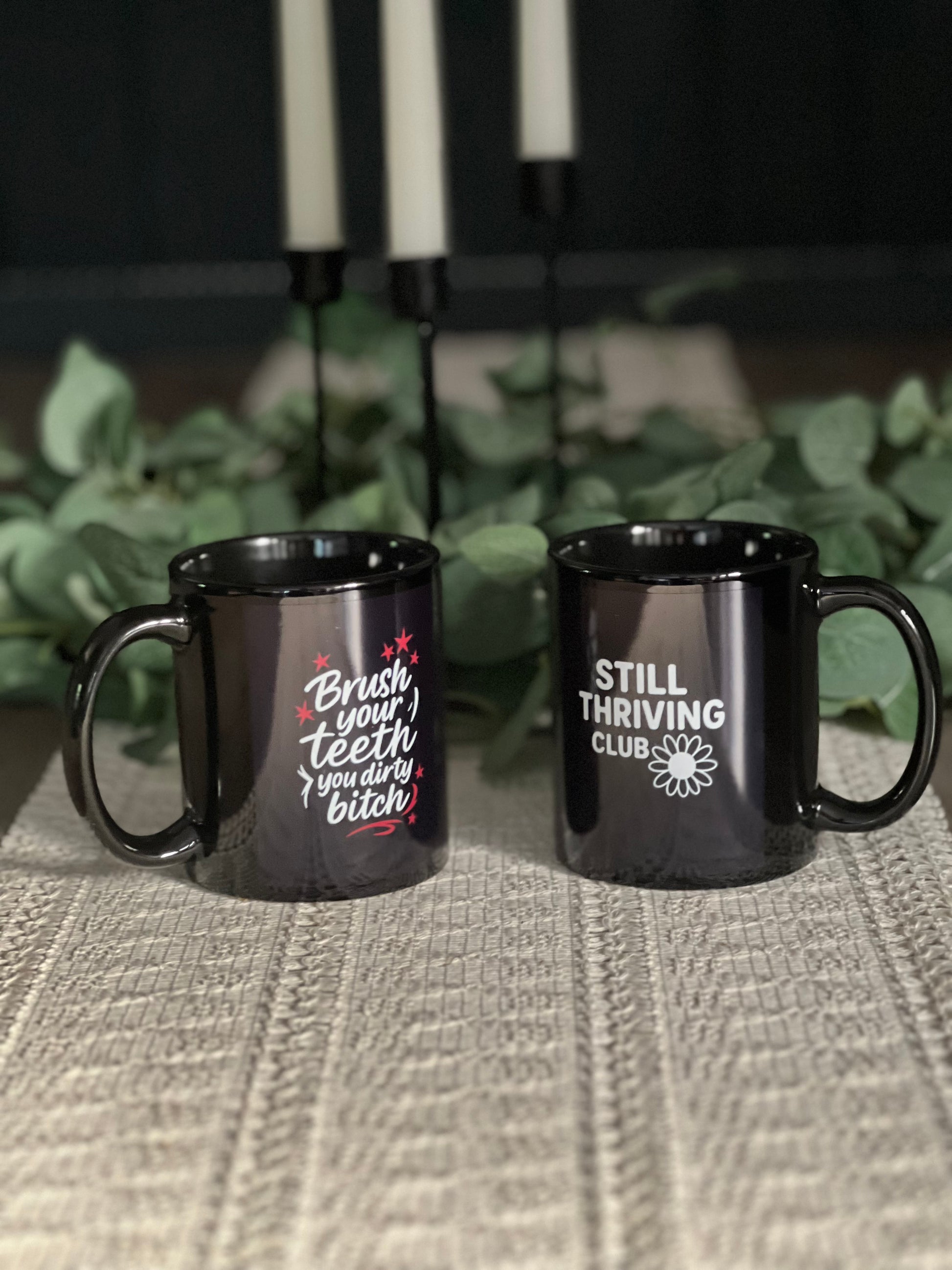 Two black ceramic mugs with Still Thriving Club designs placed on a textured surface, with green plants softly blurred in the background.