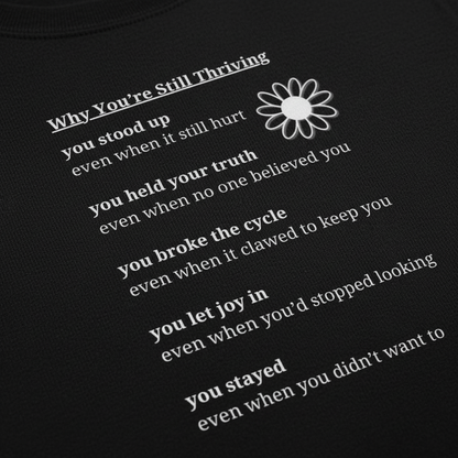 Close-up detail of black hoodie showing “Why You’re Still Thriving” affirmation text and Still Thriving Club flower logo printed on soft fabric.