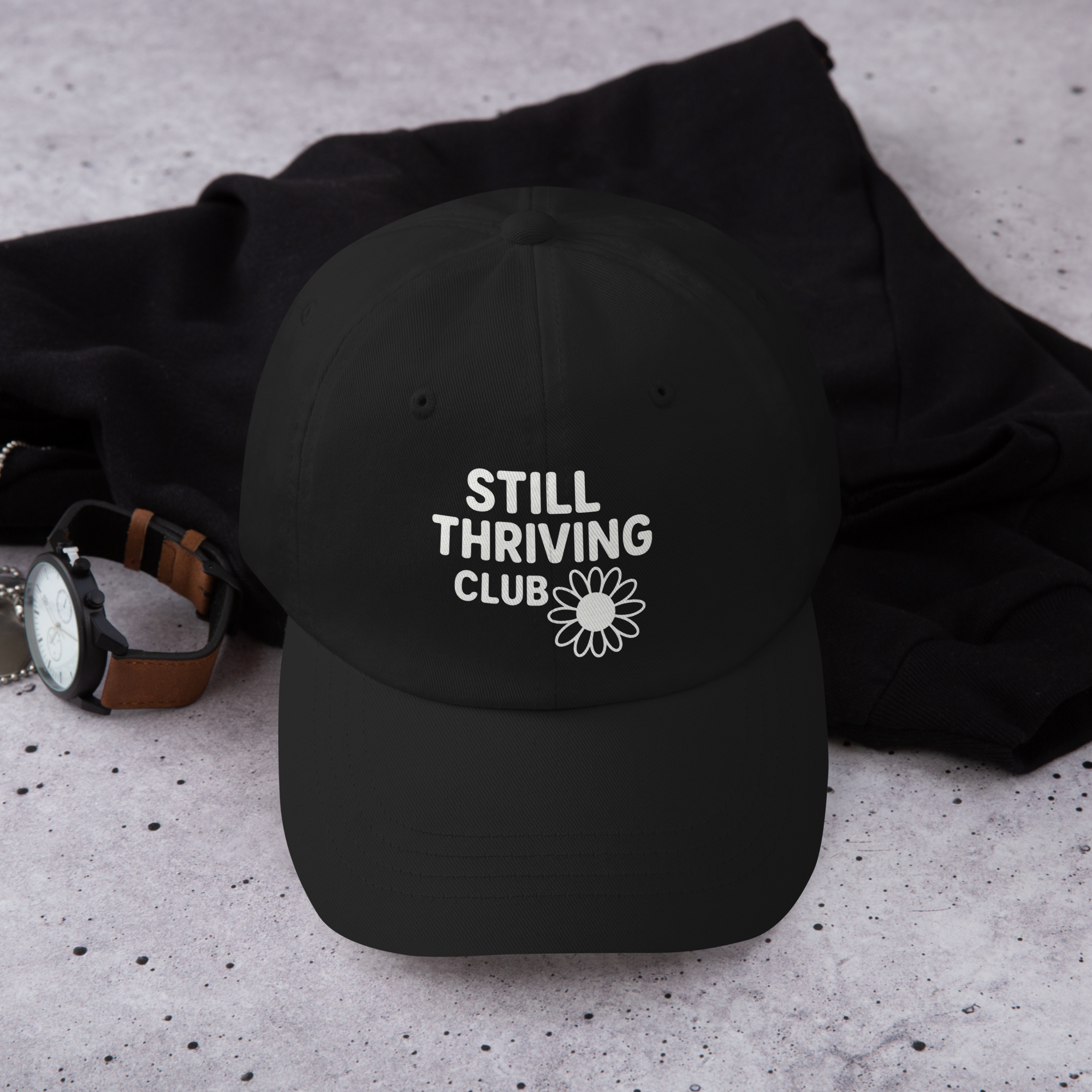Black cap with 'Still Thriving Club' text and daisy design on a textured surface.