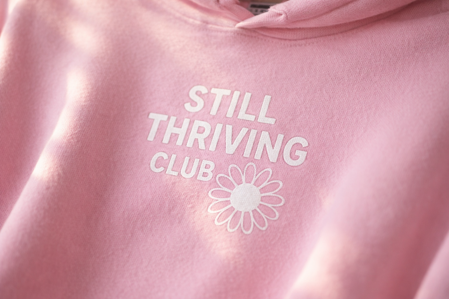 Close-up of the Still Thriving Club logo printed on a pink hoodie, highlighting the white text and flower design on soft fabric.