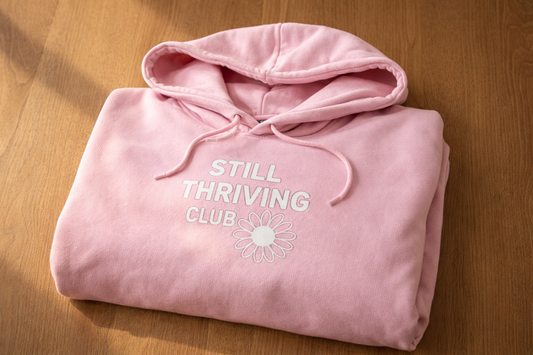 Folded pink Still Thriving Club hoodie on a wooden surface, showing the front logo and soft fabric texture in natural light.
