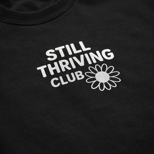 Close-up of the white Still Thriving Club logo and flower printed on black fabric, highlighting the texture of the t-shirt.
