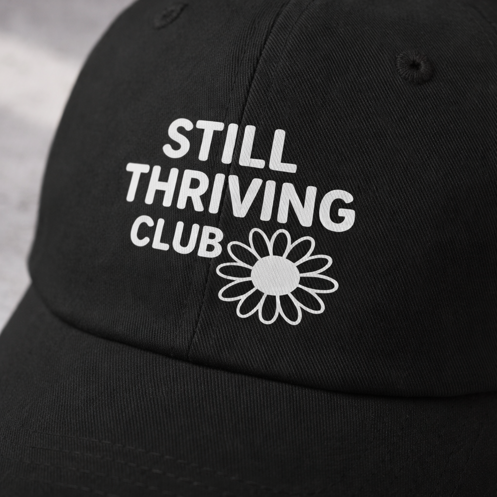 Close-up of the white Still Thriving Club logo printed on a black baseball cap, highlighting the fabric texture and stitching detail.