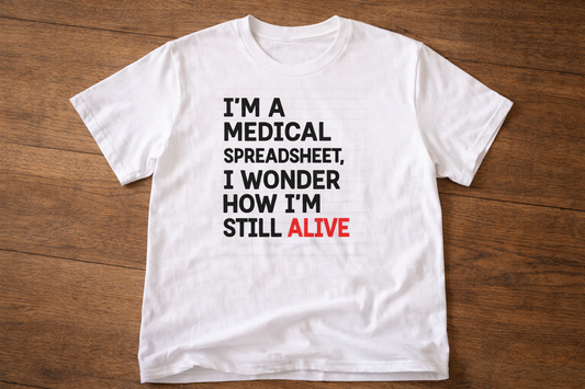 White T-shirt laid flat on a wooden surface with the text “I’m a medical spreadsheet, I wonder how I’m still alive” printed in black and red.