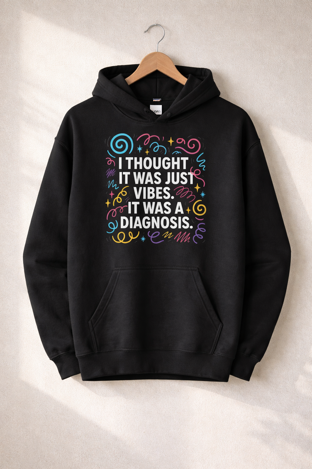 Black hoodie hanging on a wooden hanger against a neutral wall, featuring a colourful design that reads “I thought it was just vibes. It was a diagnosis.
