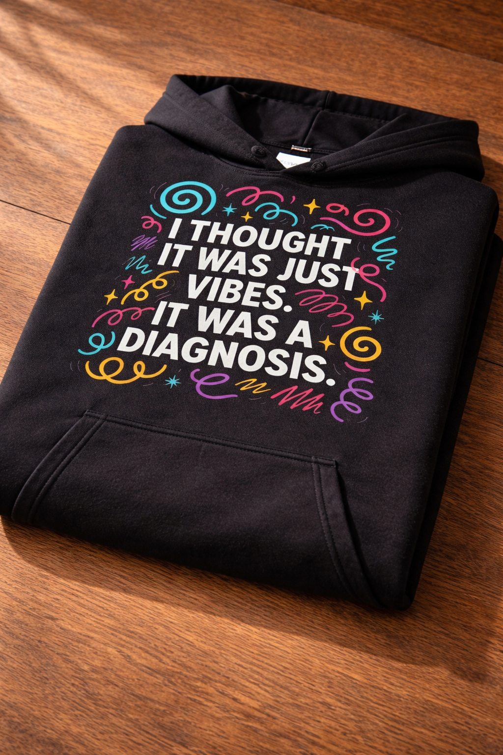 Folded black hoodie on a wooden surface, showing a close-up of the colourful “I thought it was just vibes. It was a diagnosis” design and print texture.