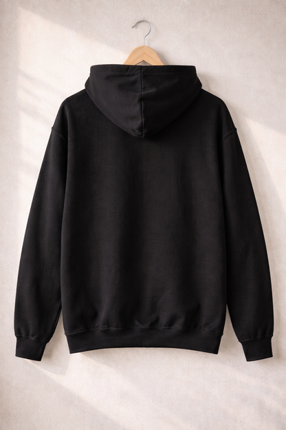 Back view of a plain black hoodie hanging on a wooden hanger against a neutral wall, showing the hood and relaxed fit.