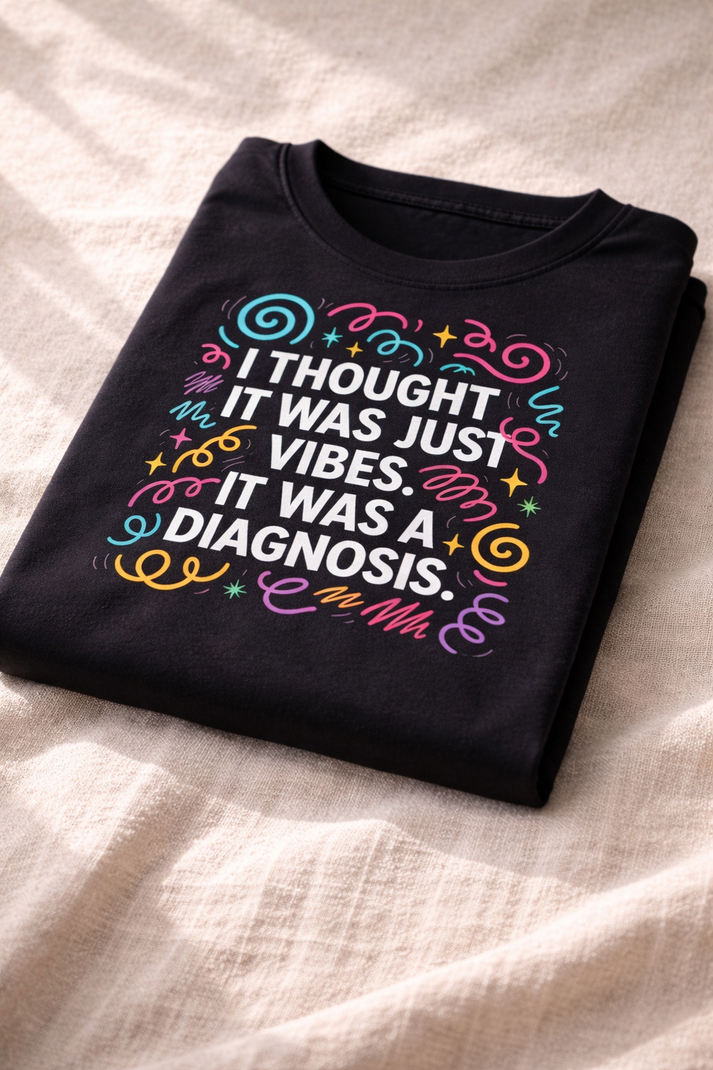 Folded black T-shirt on soft fabric showing a close-up of the colourful graphic text “I thought it was just vibes. It was a diagnosis.