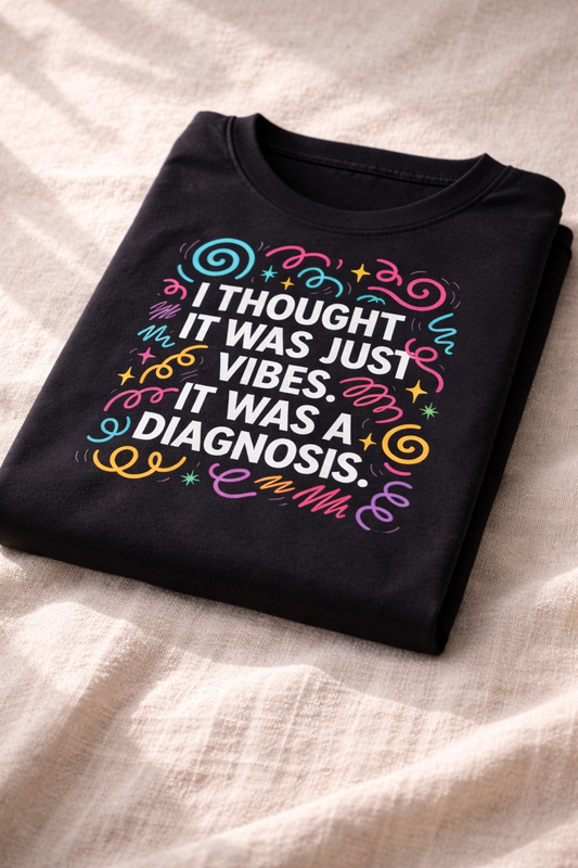 Folded black T-shirt on soft fabric showing a close-up of the colourful graphic text “I thought it was just vibes. It was a diagnosis.