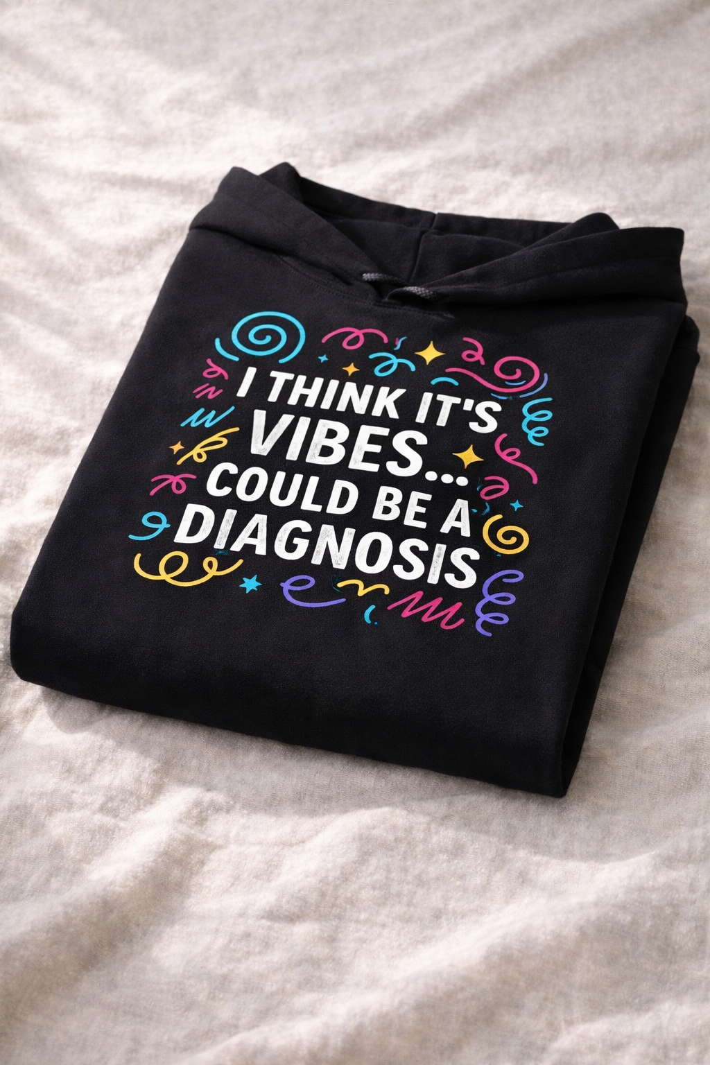 Folded black hoodie on a soft neutral surface showing the “I think it’s vibes… could be a diagnosis” design, highlighting cozy fabric and relaxed fit.