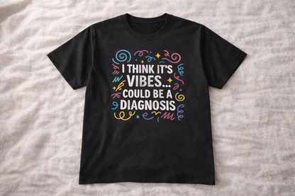 Black t-shirt laid flat on fabric background with colourful text design saying “I think it’s vibes… could be a diagnosis”.