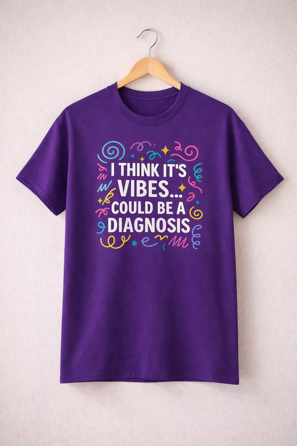 Purple t-shirt on a wooden hanger featuring the slogan “I think it’s vibes… could be a diagnosis” in colourful lettering.