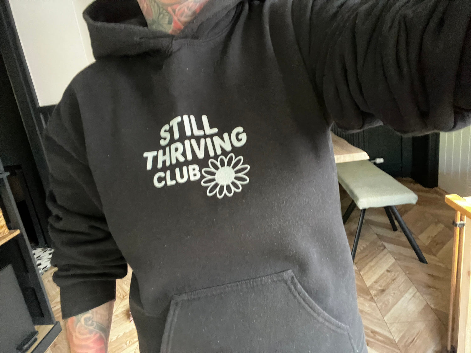 Customer wearing black Still Thriving Club hoodie with white daisy logo, shown from chest level in indoor setting