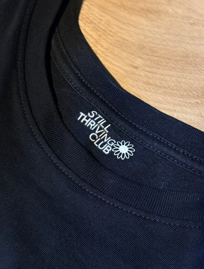 Close-up of a black t-shirt neckline showing no tags just a printed 'Still Thriving Club' logo on a wooden surface