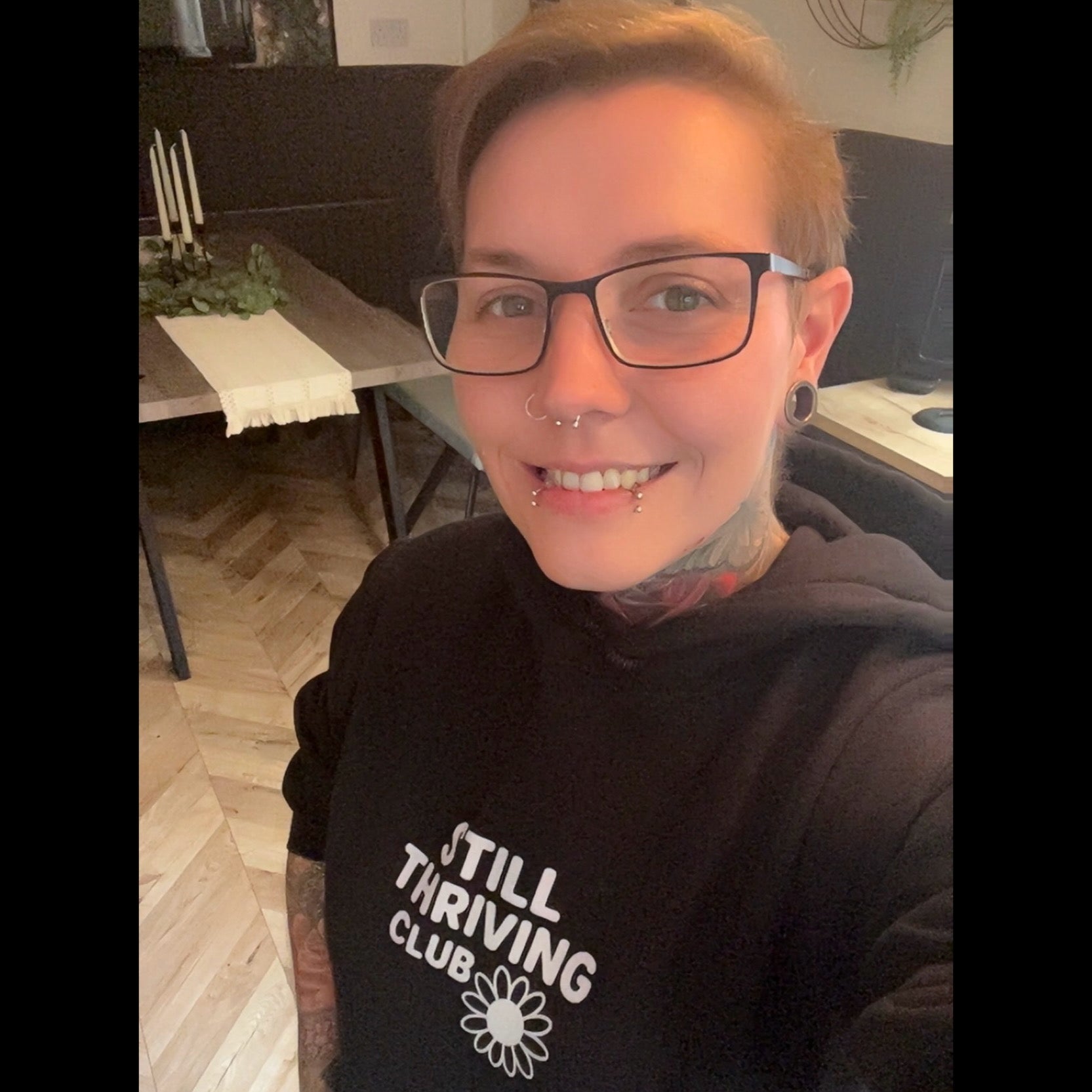 Still Thriving Club founder wearing black hoodie with white text reading 'Still Thriving Club' and daisy logo, smiling at camera