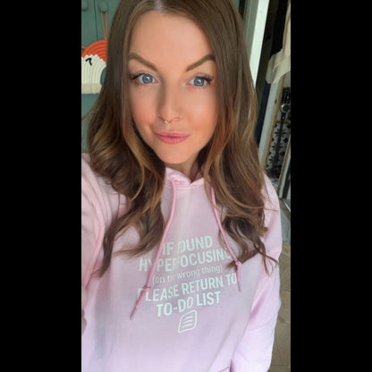 Customer wearing light pink hoodie with white text reading 'If found hyperfocusing on the wrong thing, please return to to-do list'