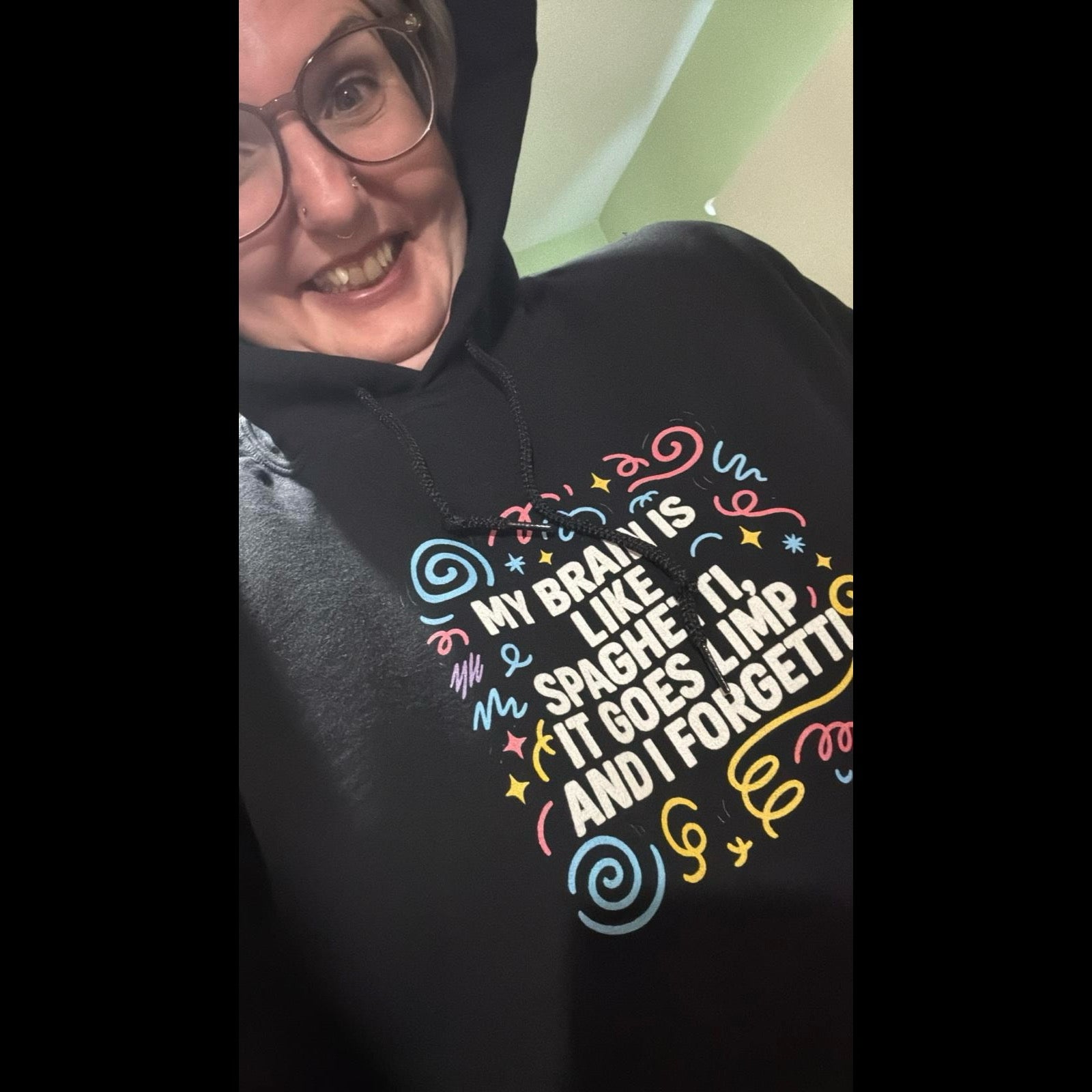 Happy customer wearing black hoodie with colorful text reading 'My brain is like spaghetti, it goes limp and I forgetti' surrounded by playful swirls and stars, smiling at camera
