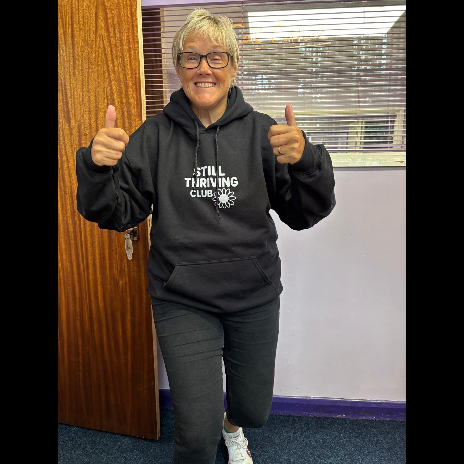 Happy customer wearing black Still Thriving Club hoodie with daisy logo, giving double thumbs up with a big smile