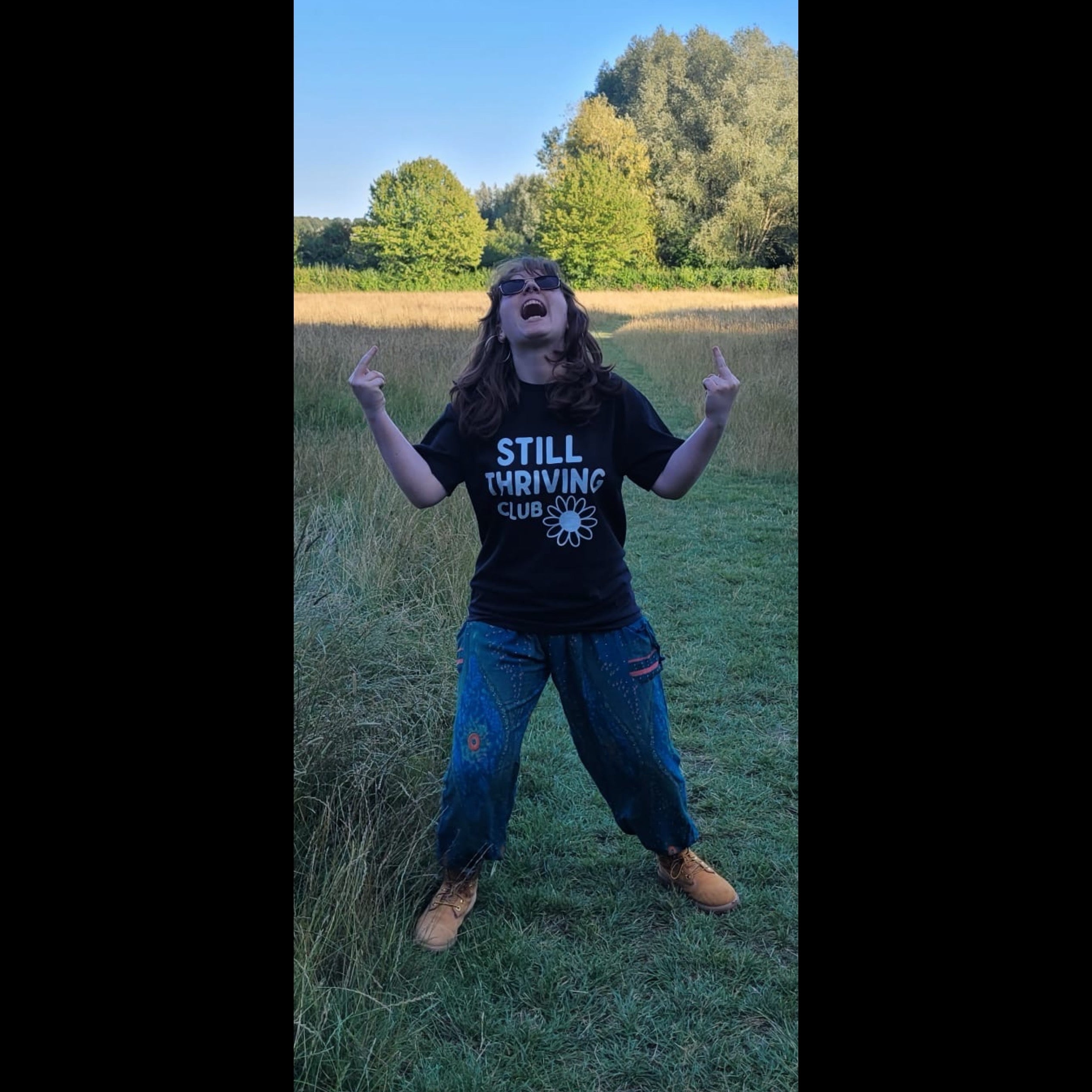 Person wearing black Still Thriving Club t-shirt with daisy logo, standing outdoors in a field with arms raised and middle fingers up, celebrating with joy