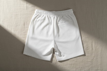 Back view of white unisex cotton shorts with elastic waistband and rear pocket, photographed flat in soft natural light