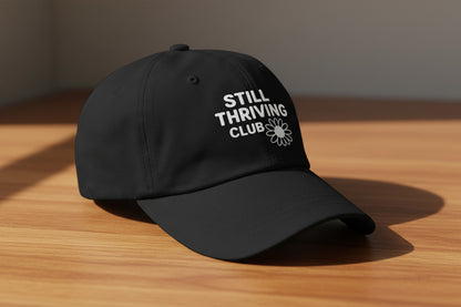 Black Still Thriving Club baseball cap resting on a wooden surface, photographed at a slight angle with soft natural light and the white logo visible on the front.