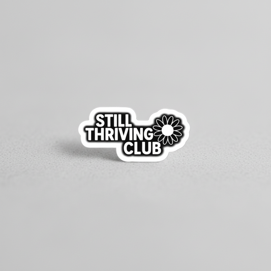 Black and white Still Thriving Club logo sticker photographed on a neutral light grey surface, showing the full sticker shape and design clearly.