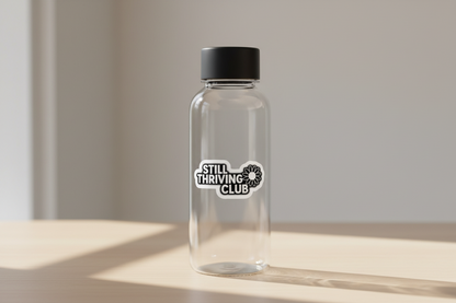Still Thriving Club logo sticker applied to a clear reusable water bottle with a black lid, resting on a light wooden surface in natural light.