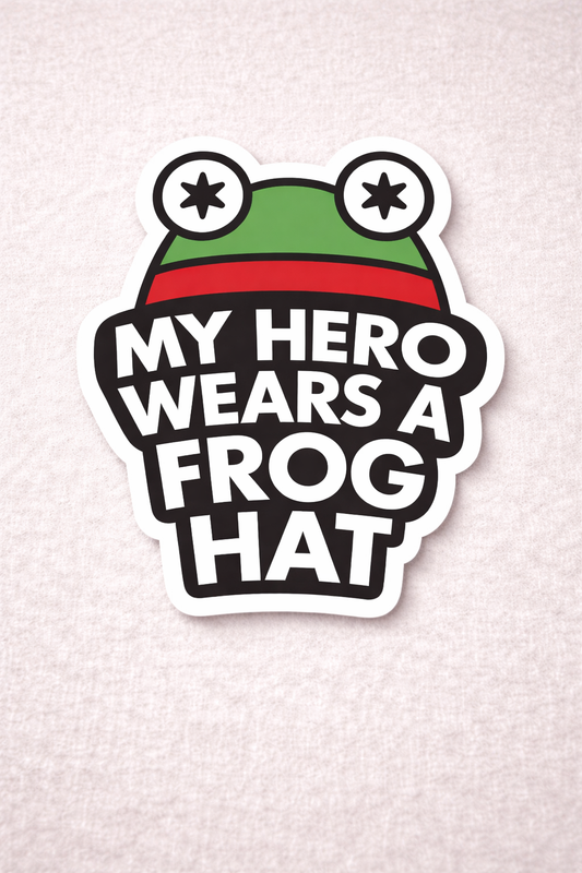Kiss cut vinyl sticker reading ‘My Hero Wears a Frog Hat’ with a green frog illustration, photographed on a neutral fabric background.