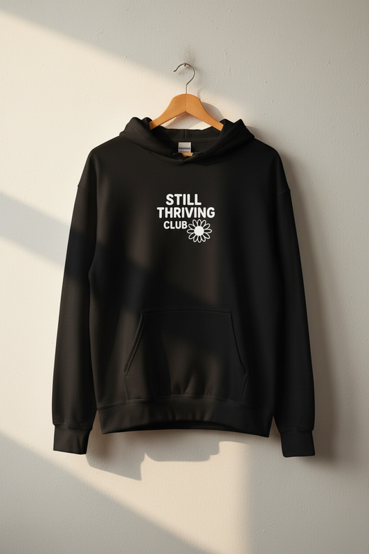Black hoodie with ‘Still Thriving Club’ logo hanging on a wooden hanger against a light wall in natural sunlight.