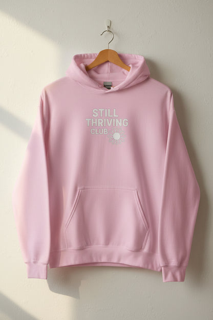Light pink Still Thriving Club hoodie hanging on a wooden hanger against a neutral wall, featuring a subtle white logo on the front in soft natural light.