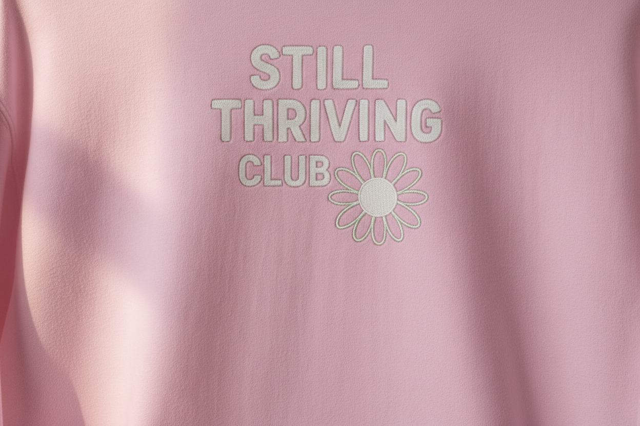 Close-up of pink hoodie fabric showing the ‘Still Thriving Club’ logo and flower emblem in soft natural light, highlighting print detail and texture.