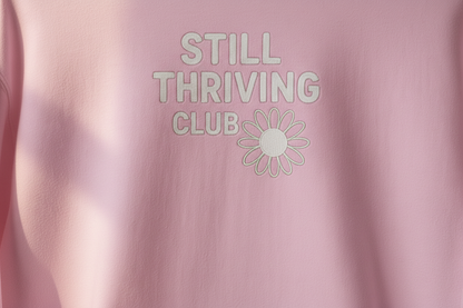 Close-up of pink hoodie fabric showing the ‘Still Thriving Club’ logo and flower emblem in soft natural light, highlighting print detail and texture.